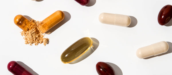 Assorted supplement capsules, including soft gels and powdered capsules, arranged on a clean white background with one capsule opened to reveal its contents.